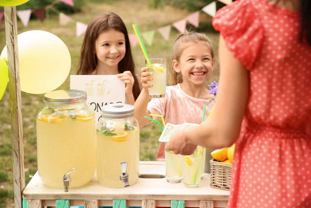 Two girls at a lemonade stand smiling. One is raising a glass of lemonade while a woman holds out a dollar bill.
