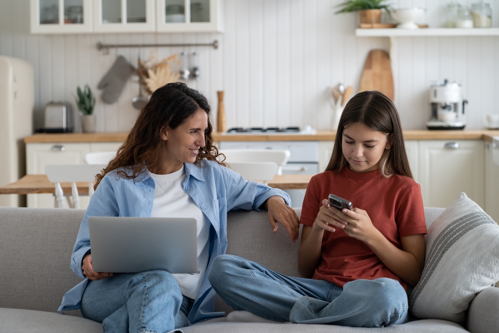 Mother and teenaged daughter sitting on a couch. Mother has a laptop and is smiling at daughter who is looking at her smartphone.